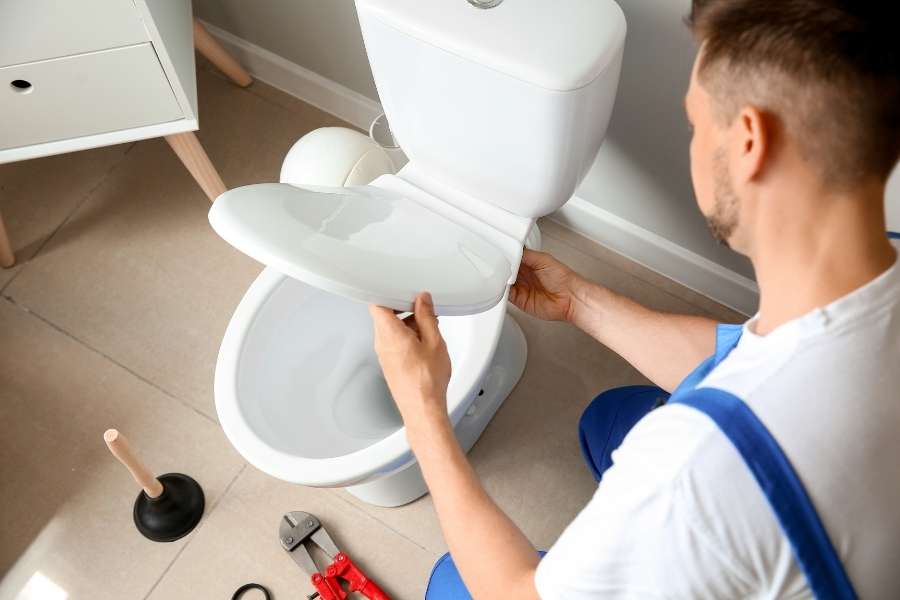Plombier en salopette bleue fixant l'abattant lors de l'installation de ses WC blancs sur carrelage beige.