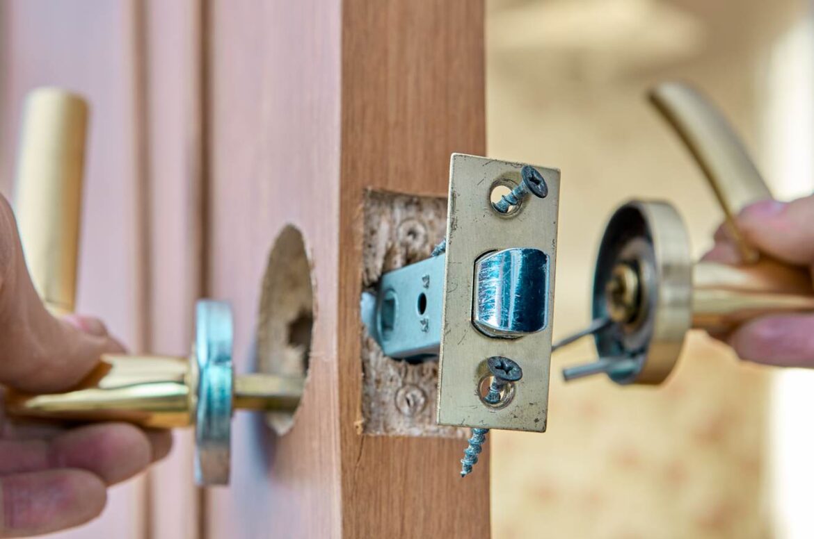 Gros plan montrant la pose du mécanisme d'une serrure sur une porte d'intérieur en bois. Un technicien tient les poignées dorées de chaque côté tandis que le loquet est vissé dans la tranche. Une illustration précise du travail réalisé pour installer verrou de blocage supplementaire serrurier paris.