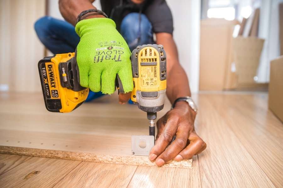 Un artisan portant un gant vert manipule une visseuse sans fil jaune pour fixer un élément sur une planche en bois.